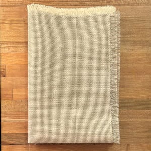 May include: A beige piece of Aida cloth with a fringed edge. The cloth is woven with a grid pattern.