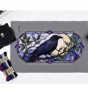 Peut inclure: Une pièce de tissu gris avec un motif au point de croix représentant un corbeau noir perché sur une branche devant une pleine lune, encadré par une bordure style vitrail aux tons violets et bleus. Le texte www.stitchingfairycrafts@etsy.com est visible.
