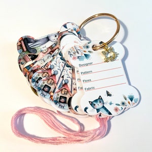 Può includere: Una carta bianca a forma di osso con un'illustrazione di un gatto e accenti floreali. La carta ha righe vuote per "Designer", "Pattern", "Floss" e "Fabric". Un anello dorato con un piccolo ciondolo a forma di gatto è attaccato alla carta.