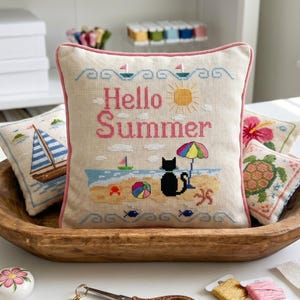 May include: A decorative pillow with the words "Hello Summer" embroidered in pink. The design includes a beach scene with a black cat, sun, umbrella, and sailboats. Other pillows with nautical and floral themes are also visible.