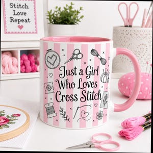 May include: A white and pink striped mug with the text "Just a Girl Who Loves Cross Stitch." The mug features cross-stitch-themed illustrations and a pink handle. Other craft supplies are in the background.