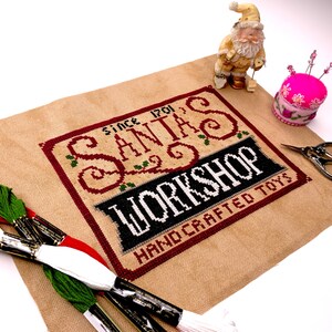 May include: A cross-stitch sampler with the text "SANTA'S WORKSHOP HAND CRAFTED TOYS SINCE 1701" in red, black, and white. The sampler is on a tan fabric, with green, red, and white embroidery floss, a Santa figurine, and a pink pincushion.
