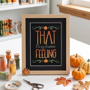 Puede incluir: Un bordado en punto de cruz enmarcado sobre tela negra con el texto en naranja y amarillo THAT Cozy Season FEELING, decorado con pequeñas calabazas naranjas y enredaderas verdes, en un marco de madera clara con paspartú blanco.