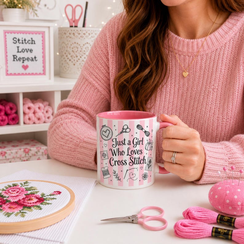 May include: A pink and white striped mug with the text "Just a Girl Who Loves Cross Stitch" and cross-stitch-themed illustrations. The mug is held near a cross-stitch project, pink thread, and scissors.