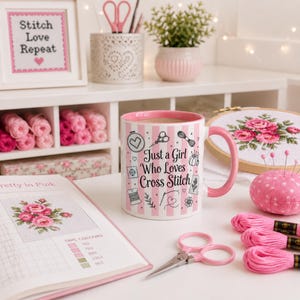 May include: A pink and white striped mug with the text "Just a Girl Who Loves Cross Stitch" sits on a white table, surrounded by cross-stitch supplies. A framed cross-stitch piece says "Stitch Love Repeat". Other items include pink thread, scissors, and a needle cushion.