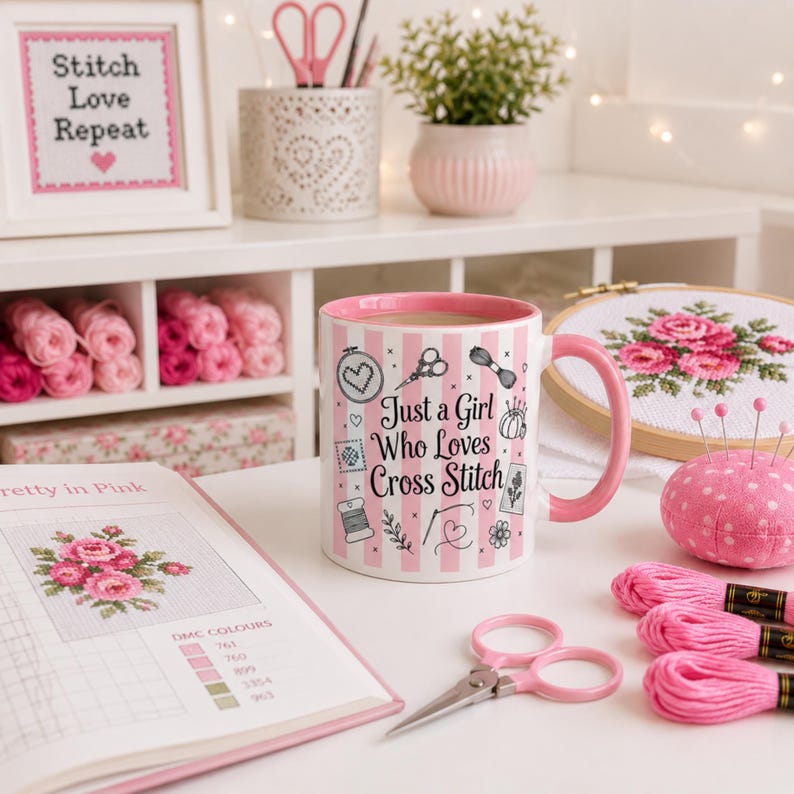 May include: A pink and white striped mug with the text "Just a Girl Who Loves Cross Stitch" sits on a white table, surrounded by cross-stitch supplies. A framed cross-stitch piece says "Stitch Love Repeat". Other items include pink thread, scissors, and a needle cushion.