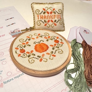 Puede incluir: Un kit de bordado de punto de cruz con un diseño de calabaza en un bastidor, una pequeña almohada con la palabra "THANKFUL" y madejas de hilo verde y marrón. El kit incluye un patrón y los materiales necesarios.