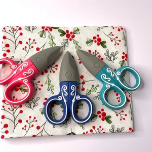 Può includere: Tre paia di piccole forbici colorate con manici decorativi. Le forbici sono su un tessuto bianco con un motivo floreale rosso e verde.