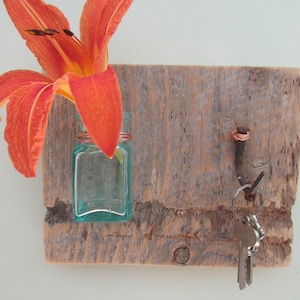 May include: A rustic wooden key holder with a small glass bottle holding an orange lily. The holder has a nail with a copper wire wrap and a hook holding keys. The wood has a natural, weathered appearance.