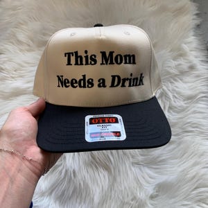 May include: A beige baseball cap with a black brim featuring the embroidered black text "This Mom Needs a Drink" on the front panel. A rectangular sticker with the brand name "OTTO" and "Classic Fit" is centered on the black brim.