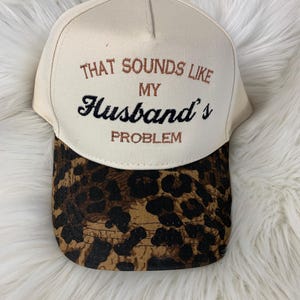 Può includere: Un cappellino da baseball beige con visiera leopardata. Il cappellino presenta la scritta ricamata "THAT SOUNDS LIKE MY Husband's PROBLEM" in filo marrone e nero.
