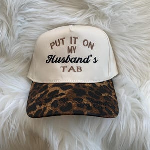 Op de afbeelding: Beige baseballpet met een luipaardprint klep. De pet heeft de geborduurde tekst "PUT IT ON MY Husband's TAB" in bruine en zwarte draad. Een casual accessoire.