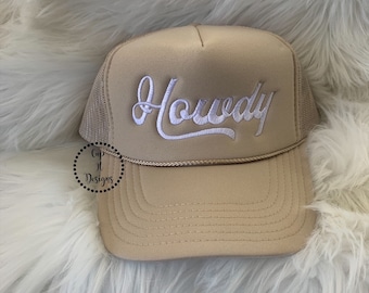 Op de afbeelding: Een beige trucker-pet met een wit geborduurd "Howdy" op de voorkant.
