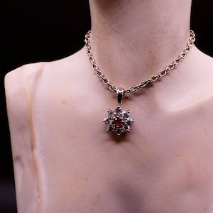 May include: A silver chain necklace with a pendant featuring a red gemstone surrounded by smaller clear gemstones in a flower shape.