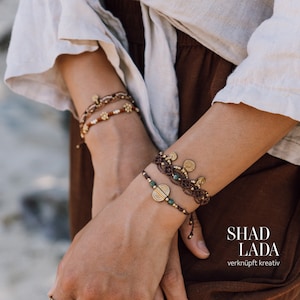 Könnte beinhalten: Das Handgelenk einer Frau ist mit vier Armbändern geschmückt. Die Armbänder sind aus braunem Kordel gefertigt und haben goldene und grüne Akzente. Die Armbänder sind übereinander gelegt und erzeugen so einen geschichteten Look. Der Text "SHAD LADA" und "verknüpft kreativ" ist in der rechten unteren Ecke des Bildes zu sehen.