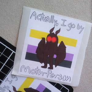 Könnte beinhalten: Ein Aufkleber mit einer Cartoon-Mottenperson mit roten Augen auf einem Hintergrund der Genderqueer-Pride-Flagge. Der Text "Actually, I go by Moth-Person" steht auf dem Aufkleber.