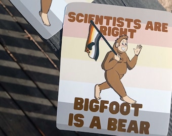 Könnte beinhalten: Zwei Aufkleber mit einem Cartoon-Bigfoot, der eine Pride-Flagge hält. Der obere Aufkleber hat einen hellbraunen, weißen und rosafarbenen Hintergrund mit dem Text "Wissenschaftler haben Recht". Der untere Aufkleber hat einen hellbraunen, weißen und rosafarbenen Hintergrund mit dem Text "Bigfoot ist ein Bär".