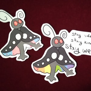 Könnte beinhalten: Zwei schwarze und weiße Cartoon-Motten-Aufkleber mit Regenbogenflügeln. Der Text auf dem Aufkleber lautet "Bleib sicher, bleib freundlich. Bleib komisch."