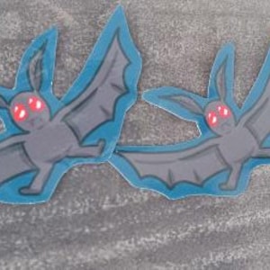 May include: Two gray bats with red eyes and blue outlines are on a gray surface. The bats are facing different directions.