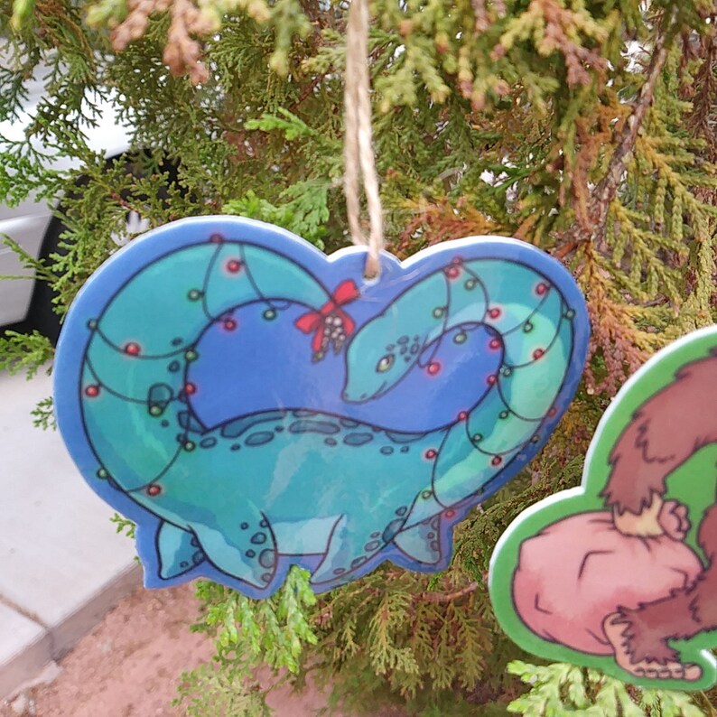 K&ouml;nnte beinhalten: Ein blauer und gr&uuml;ner herzf&ouml;rmiger Weihnachtsanh&auml;nger mit einem Cartoon-Plesiosaurus, um den sich rote und gr&uuml;ne Weihnachtslichter winden.