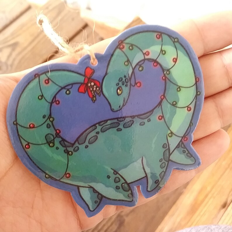 K&ouml;nnte beinhalten: Ein gr&uuml;ner und blauer Weihnachtsbaumschmuck in Herzform mit einem gr&uuml;nen und roten Dinosaurier, der sich um sich selbst wickelt. Der Dinosaurier hat rote und gr&uuml;ne Lichter um seinen Hals.