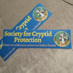 May include: Two blue and yellow bumper stickers with the text "Society for Cryptid Protection" and "Protect our wilderness, protect our hidden friends!"  The stickers feature a cartoon illustration of a cryptid in a desert landscape.