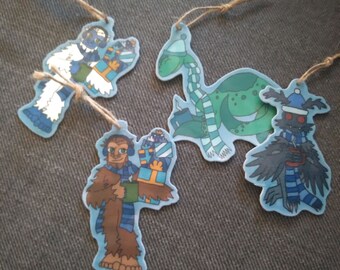 May include: Four Christmas ornaments featuring different characters. A blue yeti holding a stack of presents, a green dinosaur wearing a blue and white scarf, a brown yeti holding a stack of presents, and a black bird wearing a blue and white scarf.