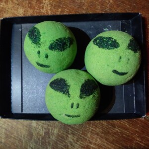 May include: Three green bath bombs with alien faces on them. The bath bombs are in a black box.