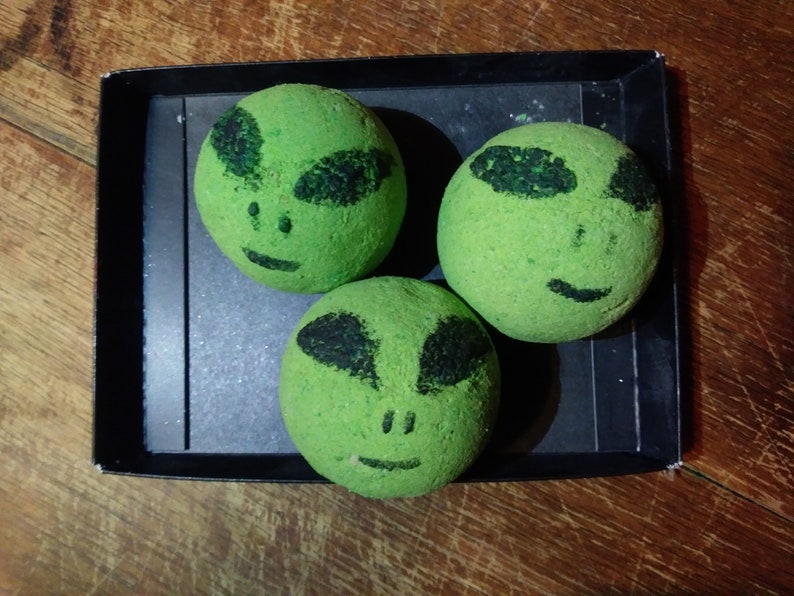 May include: Three green bath bombs with alien faces on them. The bath bombs are in a black box.