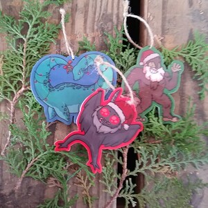 K&ouml;nnte beinhalten: Drei Weihnachtsbaumschmuck mit Comicfiguren. Ein gr&uuml;nes und blaues Wesen mit roten Lichtern, ein brauner Yeti mit Weihnachtsmannm&uuml;tze und ein schwarzes Wesen mit roten Augen und Weihnachtsmannm&uuml;tze.