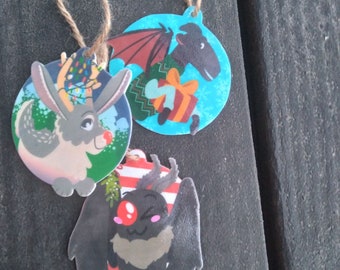 May include: Three Christmas ornaments featuring a black and white reindeer, a black bat with red eyes, and a black and white creature with wings and a red nose. The ornaments are all round and have a blue background.