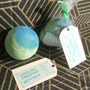 May include: A blue and green bath bomb with a glittery finish sits next to a wrapped bath bomb and two tags. The tags read "Loveland Frogman Bath Bomb" and "Rain Water Lily Grass". The bath bombs are on a black and gold geometric patterned surface.