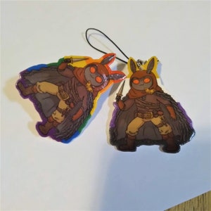 May include: Two colorful keychains featuring a cartoon character with brown fur, red eyes, and a brown hooded cloak. The character is holding a dagger in one hand and has a second dagger strapped to their leg. The keychain on the left has a rainbow background, while the keychain on the right has a purple and white background.