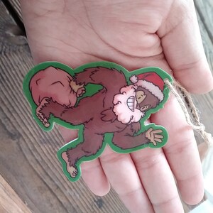 K&ouml;nnte beinhalten: Ein gr&uuml;ner Weihnachtsbaumschmuck in Form eines braunen Bigfoots, der einen roten Sack tr&auml;gt. Der Bigfoot tr&auml;gt eine rote Weihnachtsm&uuml;tze und einen wei&szlig;en Bart.