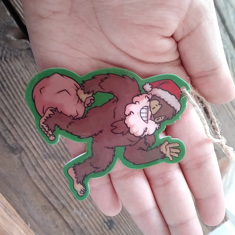 K&ouml;nnte beinhalten: Ein gr&uuml;ner Weihnachtsbaumschmuck in Form eines braunen Bigfoots, der einen roten Sack tr&auml;gt. Der Bigfoot tr&auml;gt eine rote Weihnachtsm&uuml;tze und einen wei&szlig;en Bart.