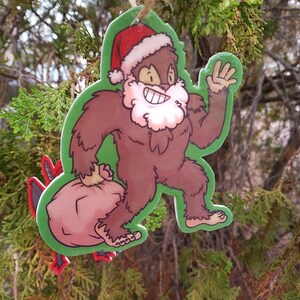 K&ouml;nnte beinhalten: Ein brauner Bigfoot, der einen roten und wei&szlig;en Weihnachtsmann-Hut tr&auml;gt und einen roten Sack tr&auml;gt. Der Bigfoot l&auml;chelt und winkt. Der Anh&auml;nger ist aus Papier und hat einen gr&uuml;nen Hintergrund.