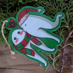 May include: A Christmas ornament shaped like a tooth wearing a Santa hat and a scarf, kissing another tooth wearing a green and red scarf. The ornament is made of white and green plastic and has a string for hanging.