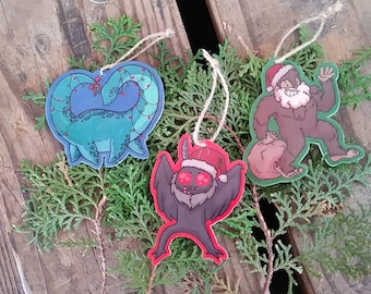 May include: Three Christmas ornaments featuring a green and blue dinosaur, a black bat with red eyes, and a brown Bigfoot wearing a red Santa hat. Each ornament has a string for hanging.