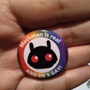 Op de afbeelding: Een regenboogkleurige knop met de tekst "Mothman is real and he's gay!" en een cartoonafbeelding van een zwarte mot met rode ogen.