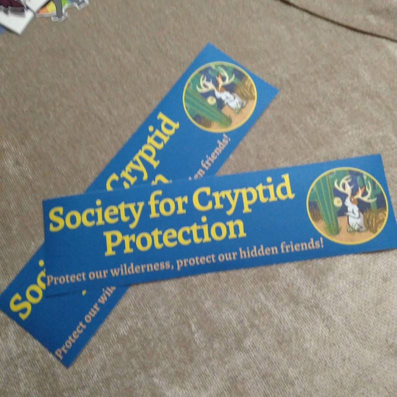 May include: Two blue and yellow stickers with the text "Society for Cryptid Protection" and a cartoon image of a cryptid. The text on the sticker reads "Protect our wilderness, protect our hidden friends!"