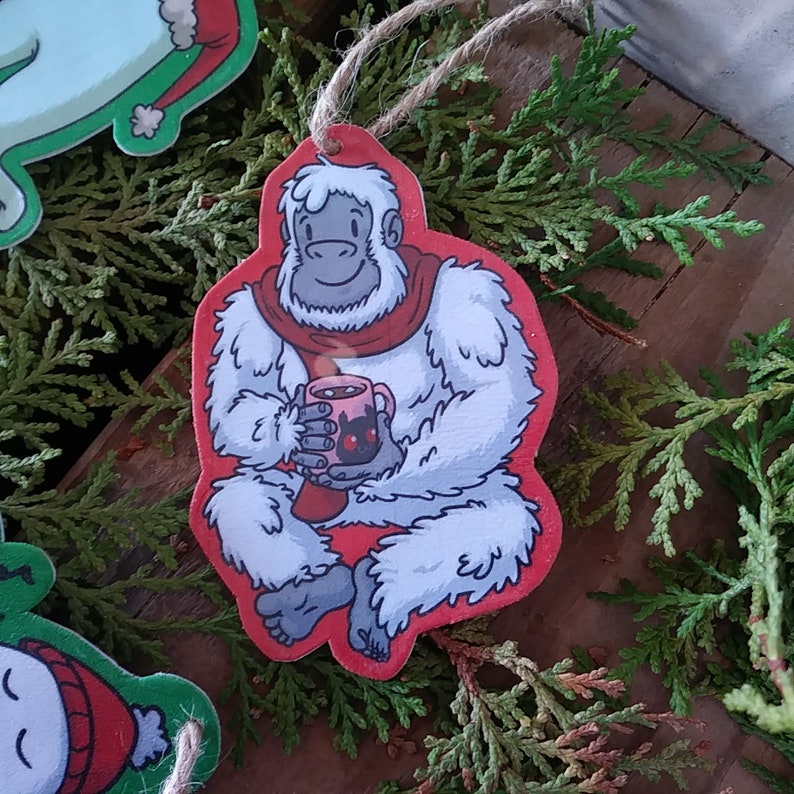 May include: A white Yeti holding a red mug with a black design. The Yeti is wearing a red scarf and is sitting on a red background.