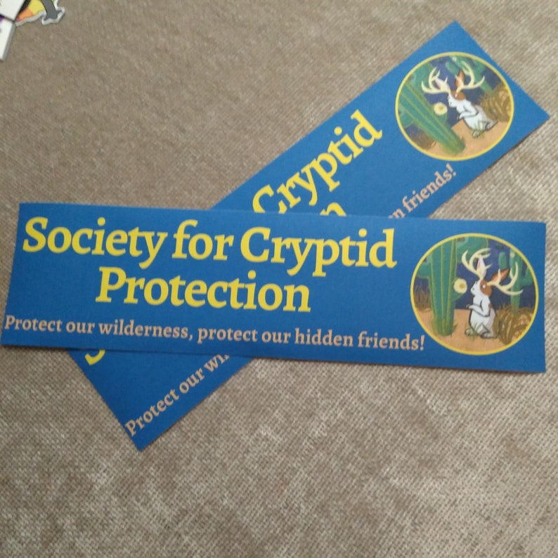 May include: Two blue and yellow bumper stickers with the text "Society for Cryptid Protection" and "Protect our wilderness, protect our hidden friends!"  The stickers feature a cartoon image of a white rabbit with antlers standing in a desert landscape.