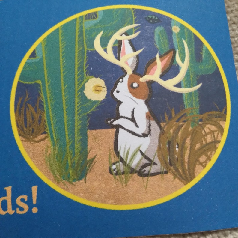 May include: A white rabbit with brown spots and antlers stands in a desert landscape. The rabbit is looking to the left, and there is a large cactus behind it. The background is a blue sky with stars.