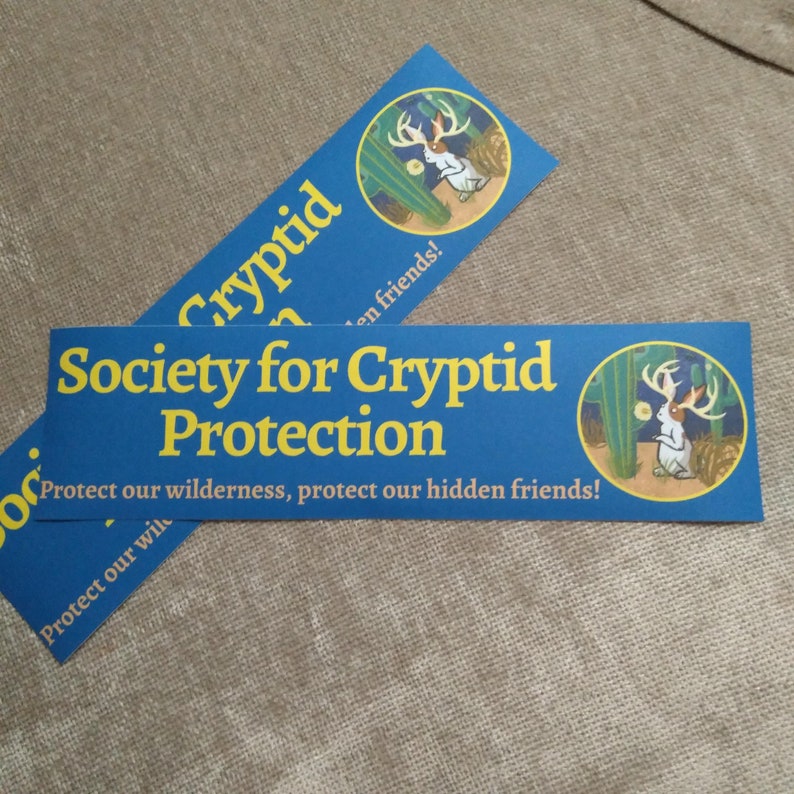 May include: Two blue and yellow bumper stickers with the text "Society for Cryptid Protection" and "Protect our wilderness, protect our hidden friends!"  The stickers feature a cartoon white rabbit with antlers in a desert landscape.