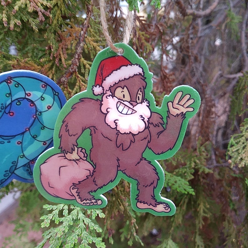 K&ouml;nnte beinhalten: Ein brauner Cartoon-Bigfoot, der einen roten Weihnachtsmann-Hut tr&auml;gt und einen braunen Sack h&auml;lt. Der Bigfoot l&auml;chelt und hat einen wei&szlig;en Bart. Der Anh&auml;nger ist aus Holz und hat eine Schnur zum Aufh&auml;ngen.