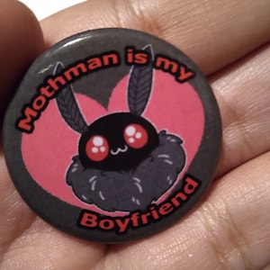 May include: A black and gray button with a pink heart and a cartoon moth with white eyes and red cheeks. The text on the button reads "Mothman is my Boyfriend".