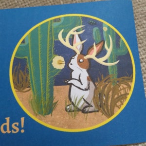 May include: A white rabbit with brown spots and antlers stands in a desert landscape. The rabbit is looking up at a cactus with a yellow flower. The background is a blue sky with stars.