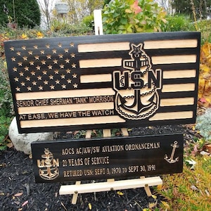 May include: A wooden sign with a black and gold American flag design. The sign features a carved US Navy anchor and the text "SENIOR CHIEF SHERMAN "TANK" MORRISON AT EASE, WE HAVE THE WATCH". A smaller wooden sign below reads "AOCS AC/AW/SW AVIATION ORDNANCEMAN USN 21 YEARS OF SERVICE RETIRED USN SEPT 3, 1970 TO SEPT 30, 1990".