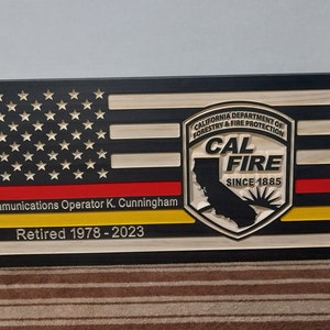 May include: A wooden plaque with a black background and a red, white, and blue American flag design. The plaque also features the California Department of Forestry and Fire Protection logo, with the text "CAL FIRE" and "SINCE 1885". The plaque has a red and yellow stripe at the bottom with the text "Communications Operator K. Cunningham Retired 1978-2023".
