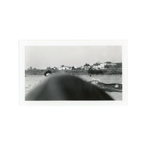 May include: Black and white photograph of a coastal landscape. The image features a sandy beach in the foreground, with a blurred dark shape obscuring part of the view. In the background, a row of buildings and houses are visible along a hillside.